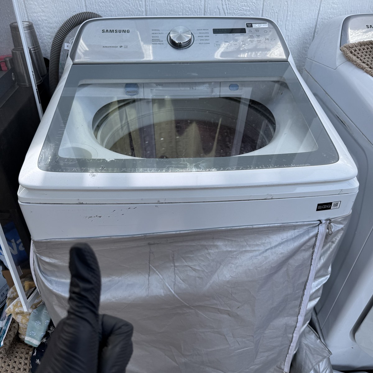 White Samsung top-load washer with a glass lid and a zip fabric cover beside a dryer in an outdoor laundry nook on a Spark service call