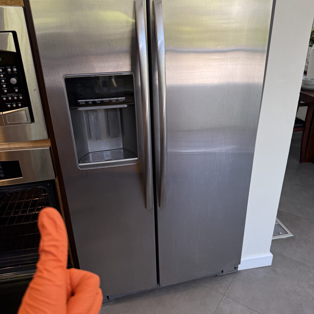 Stainless side-by-side refrigerator with a door ice and water dispenser beside a wall oven, a technician’s orange glove in frame