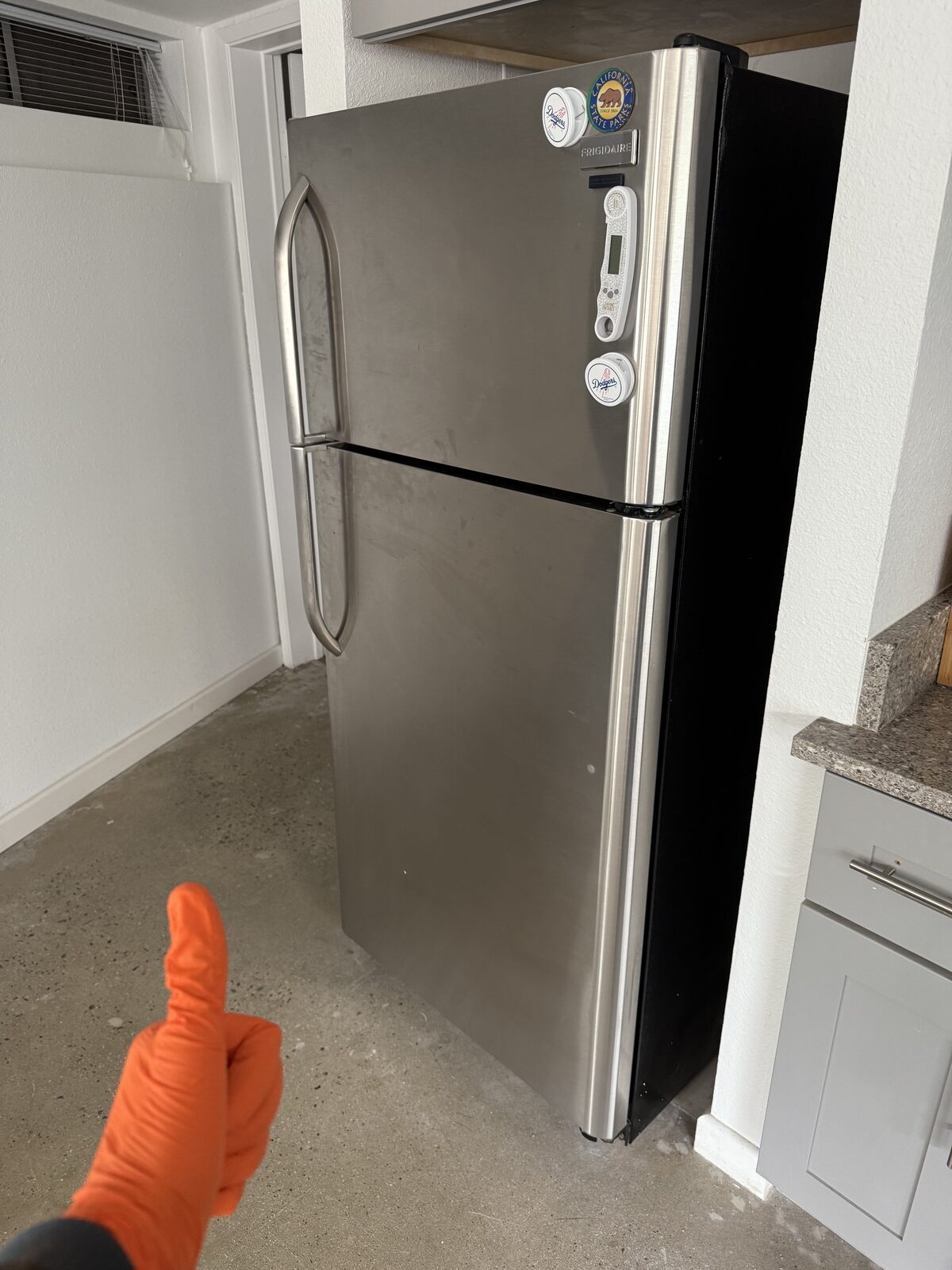 Stainless Frigidaire top-freezer refrigerator on a concrete floor beside a kitchen counter, a technician’s gloved thumbs-up in frame
